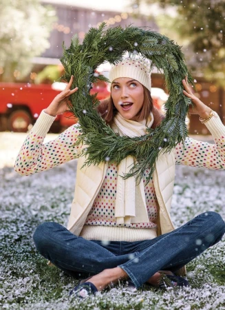 A smiling woman sits cross-legged in a snowy outdoor scene, holding a wreath up to her face, wearing a winter hat, scarf, vest, and jeans.
