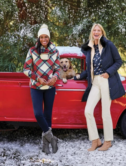 Two adult women pose next to a red truck in the snow with a dog in winter clothing, creating a festive outdoor scene.