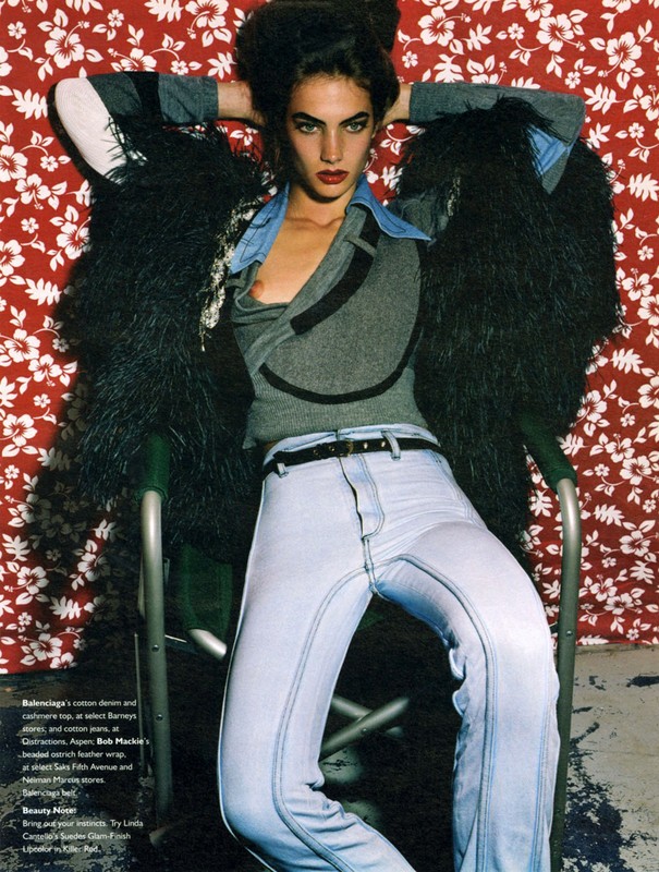 Woman in retro fashion: gray top, blue jeans, faux fur coat posing against floral backdrop in a vintage-style fashion editorial.