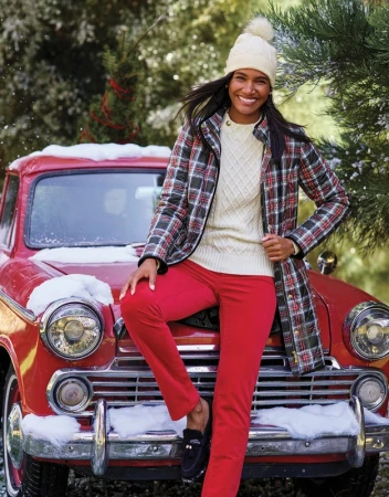 Smiling woman in plaid coat poses on red vintage car with snow, wearing red pants and a white beanie, surrounded by green foliage.
