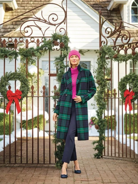Adult woman in a plaid overcoat, pink sweater, and hat stands outdoors near a decorated wrought-iron gate and brick walkway.