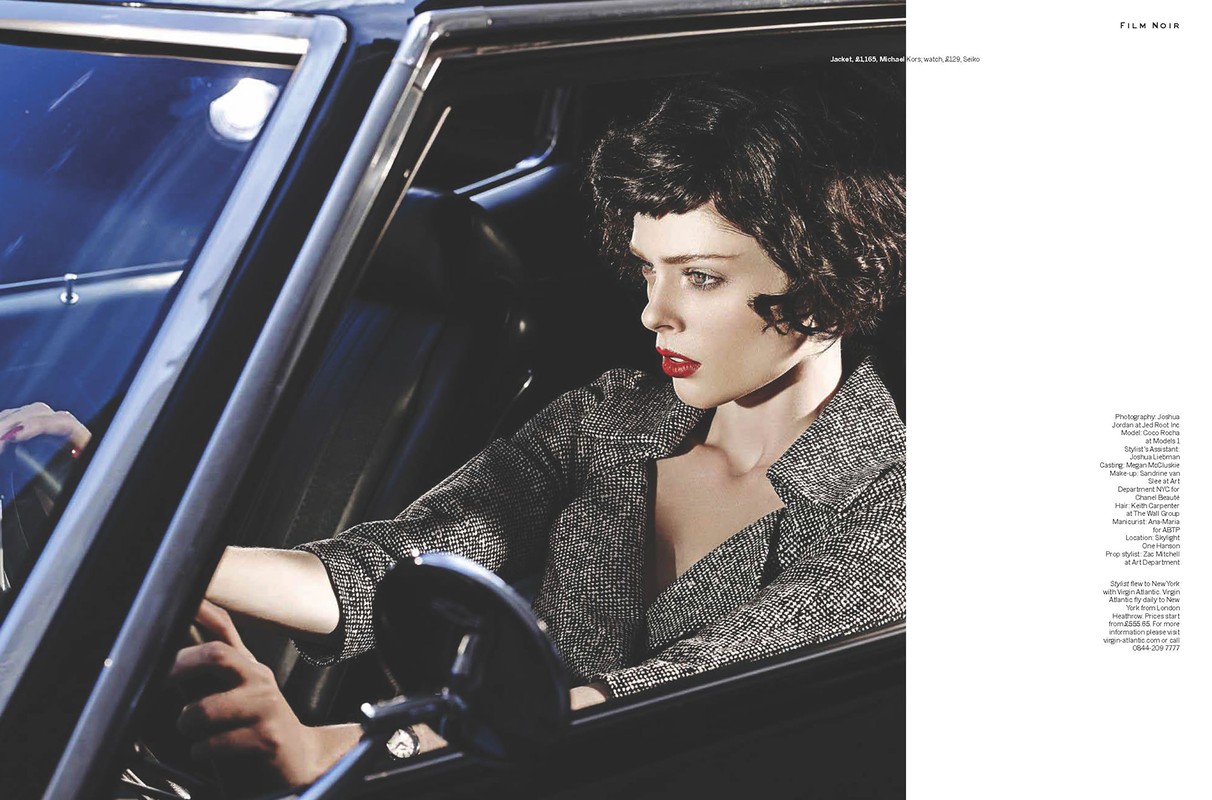 Fashion shot of a brunette woman in a tweed jacket driving a classic black car, red lipstick, moody lighting, and film noir style.
