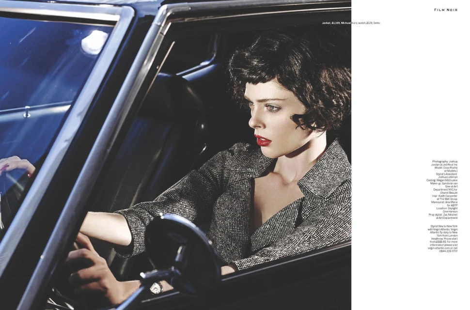 Fashion shot of a brunette woman in a tweed jacket driving a classic black car, red lipstick, moody lighting, and film noir style.