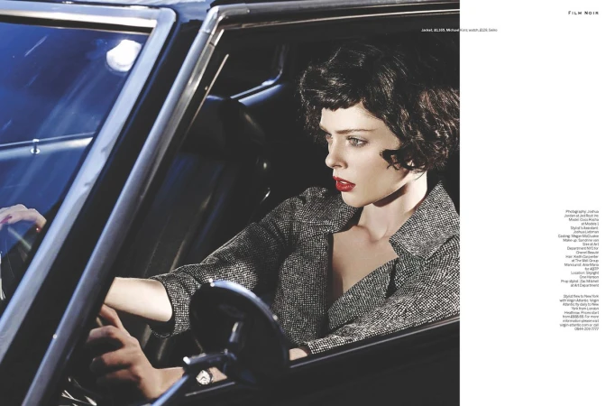 Fashion shot of a brunette woman in a tweed jacket driving a classic black car, red lipstick, moody lighting, and film noir style.