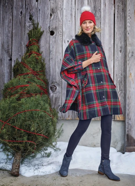 Woman in a plaid cape, red beanie, and blue boots standing next to a small Christmas tree outdoors with a wooden fence background.
