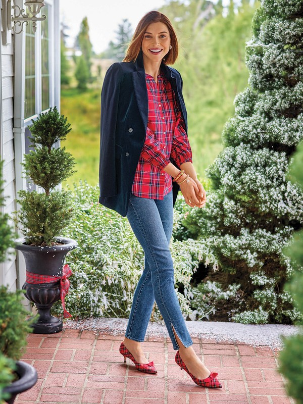 Woman wearing a red plaid top and denim jeans poses outdoors with a black blazer draped over her shoulder, smiling at the camera.