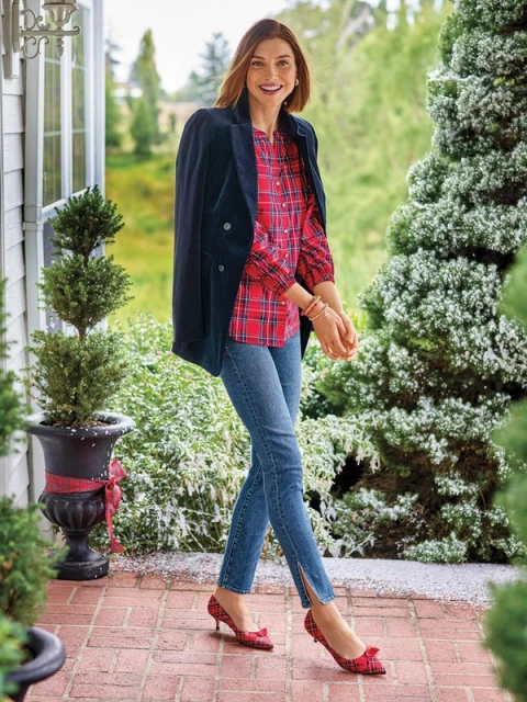 Woman wearing a red plaid top and denim jeans poses outdoors with a black blazer draped over her shoulder, smiling at the camera.