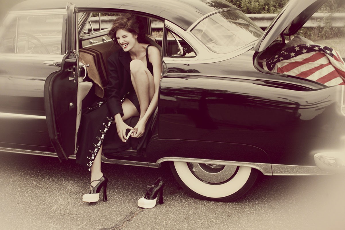 Fashion shot of a smiling woman with a vintage car, high heels, and an American flag in the trunk in sepia tones outdoors.