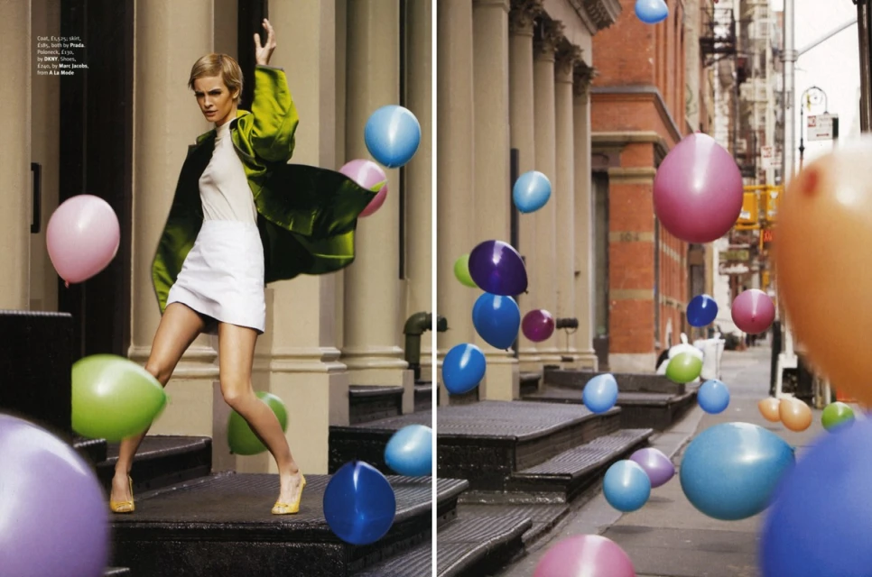 Fashion editorial featuring a woman in a green coat and white skirt surrounded by colorful balloons on an urban street with buildings.