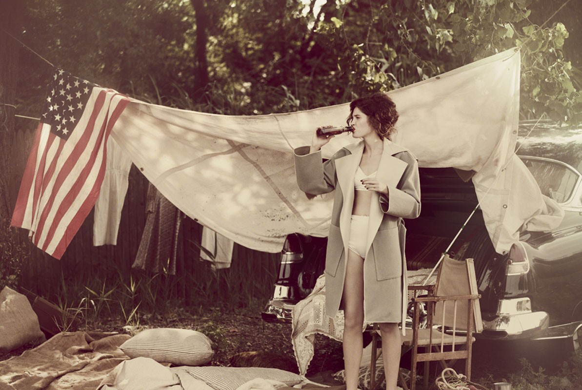Woman in trench coat drinks from bottle near USA flag draped over a vintage car in outdoor setting with a green forest backdrop.