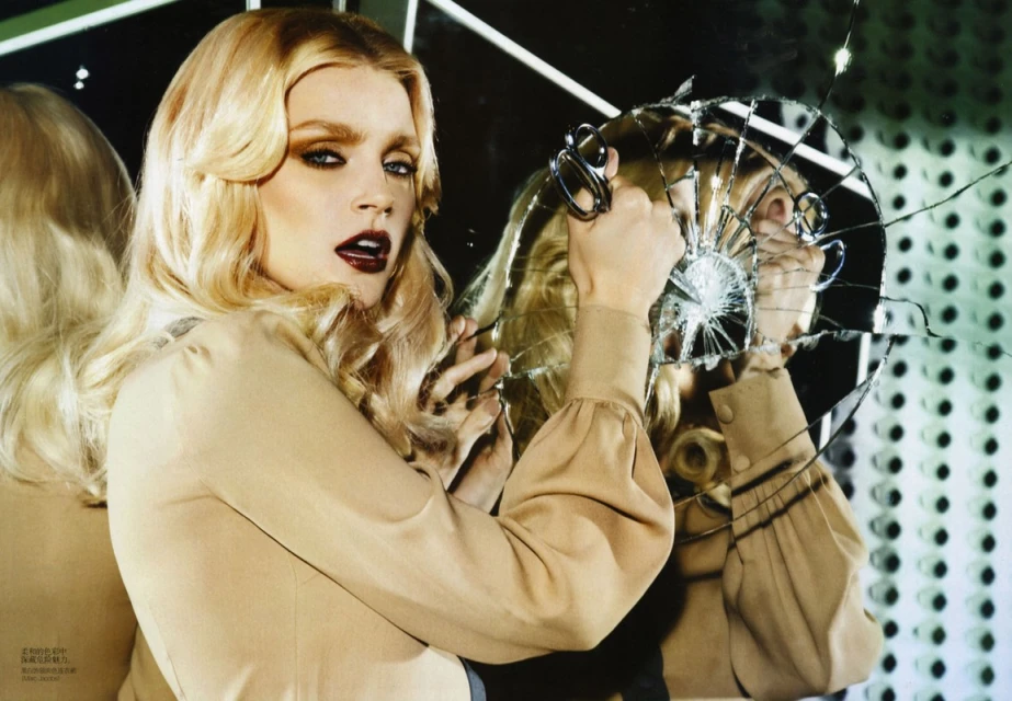 Attractive blond woman in beige blouse holding scissors against cracked mirror in studio setting with green dotted background, fashion portrait.