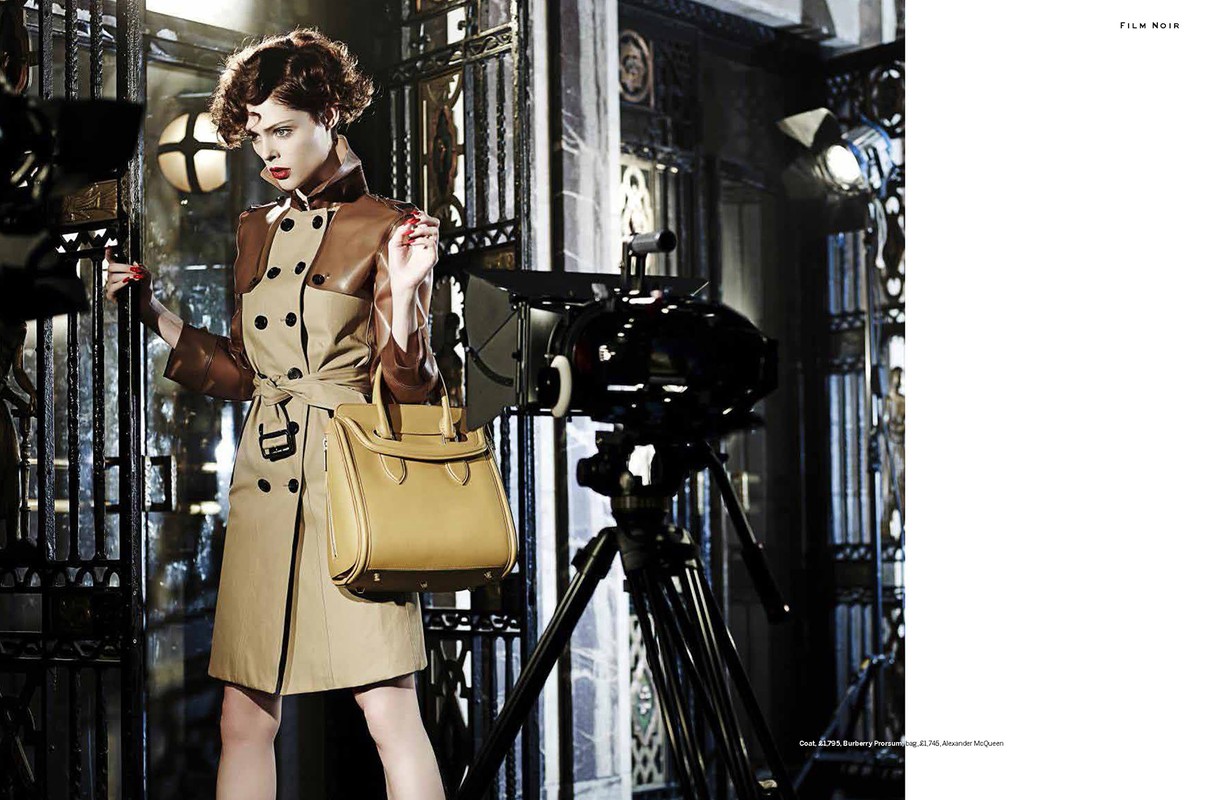 Elegant woman in trench coat holds handbag next to movie camera in a dark studio, showcasing a vintage fashion editorial.