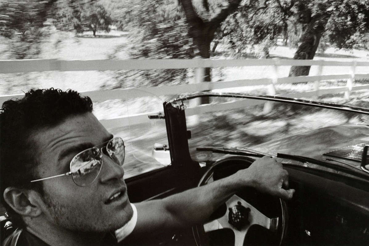 Monochrome shot of a man driving a vintage convertible with a white fence and blurred trees in the background, creating a sense of motion.