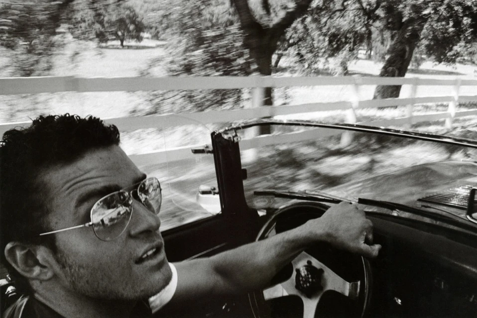 Monochrome shot of a man driving a vintage convertible with a white fence and blurred trees in the background, creating a sense of motion.
