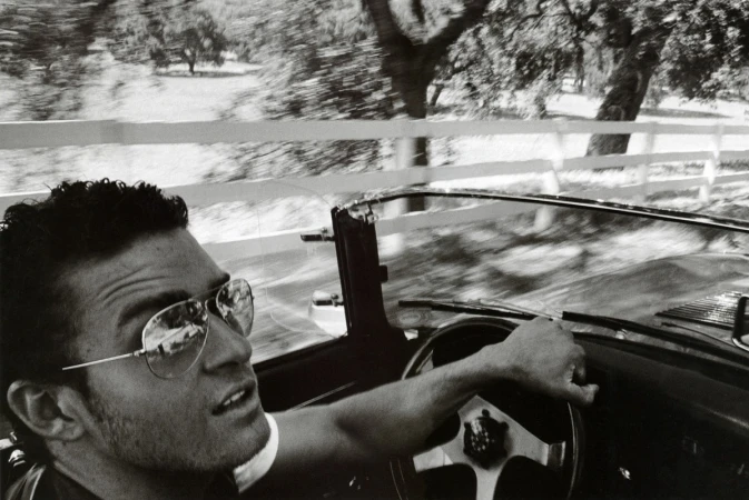 Monochrome shot of a man driving a vintage convertible with a white fence and blurred trees in the background, creating a sense of motion.