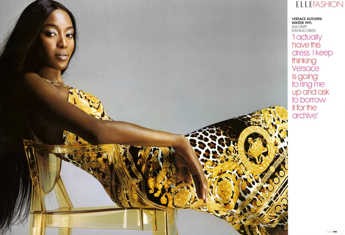 Naomi Campbell in Versace dress, laying on chair, long dark hair, gold patterned evening dress, indoor shot. Fashion editorial photo.