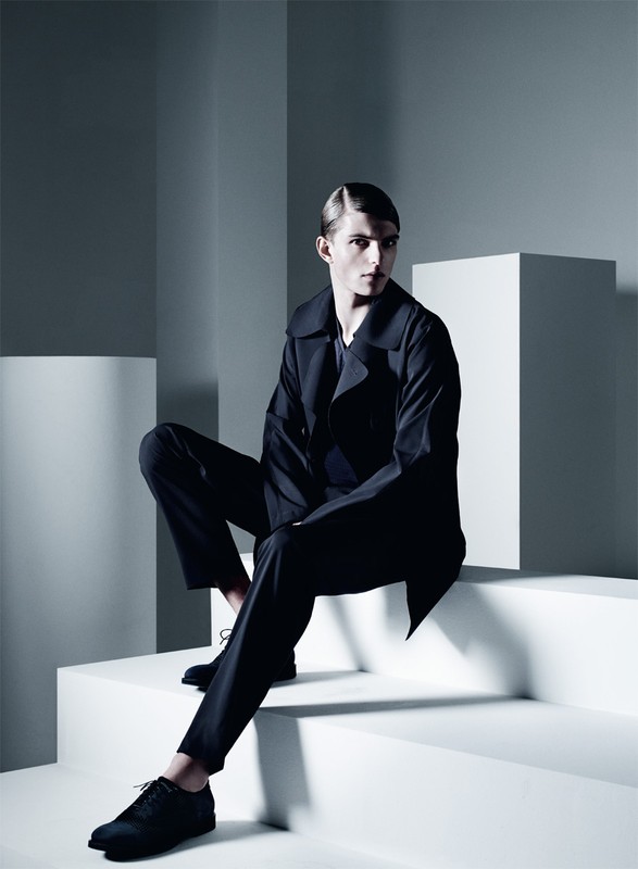 A male model in a dark suit sits on white geometric steps in a minimalist studio. Cool, neutral tones and shadows accentuate the fashion shot.
