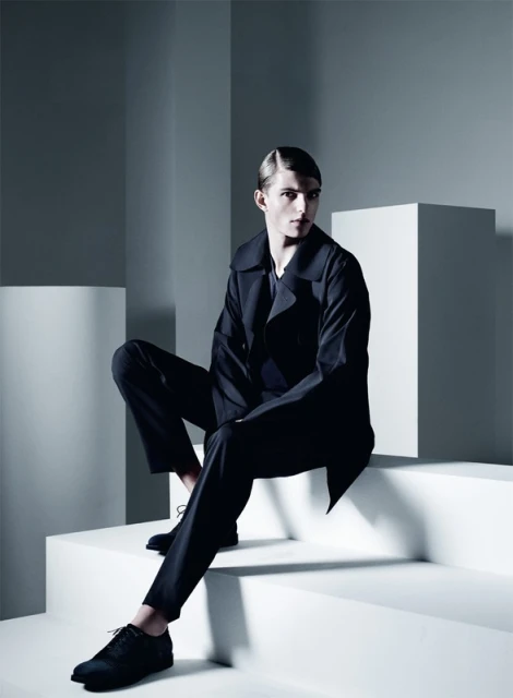 A male model in a dark suit sits on white geometric steps in a minimalist studio. Cool, neutral tones and shadows accentuate the fashion shot.