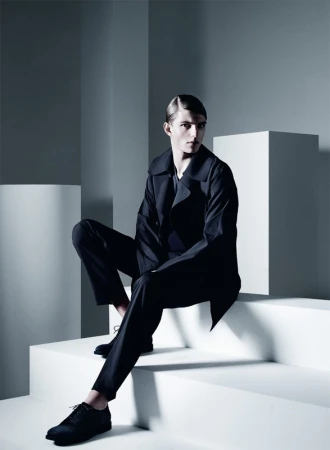 A male model in a dark suit sits on white geometric steps in a minimalist studio. Cool, neutral tones and shadows accentuate the fashion shot.