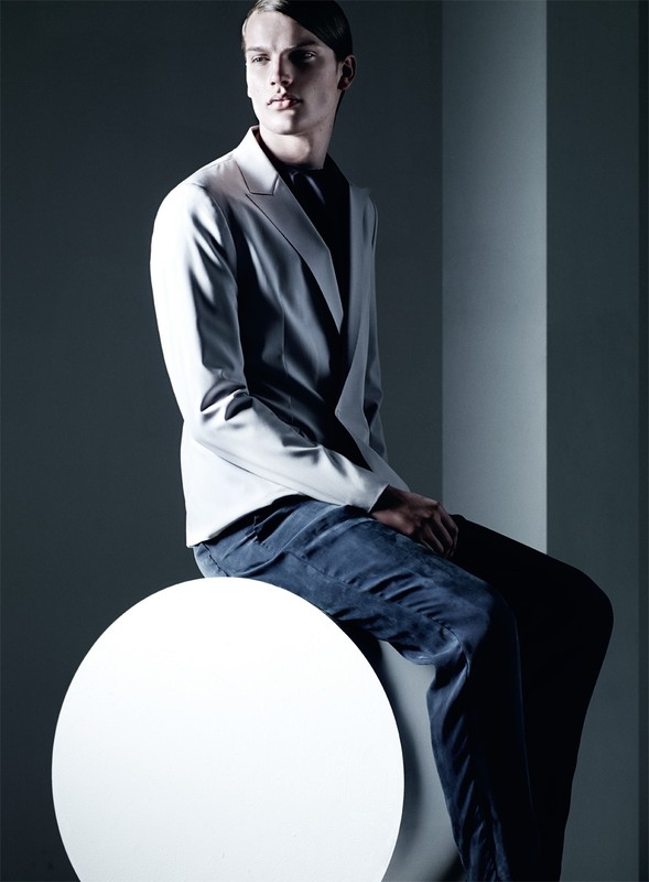 Fashion shot of a male model in a light grey suit sitting on a white sphere in a photo studio with directional lighting.
