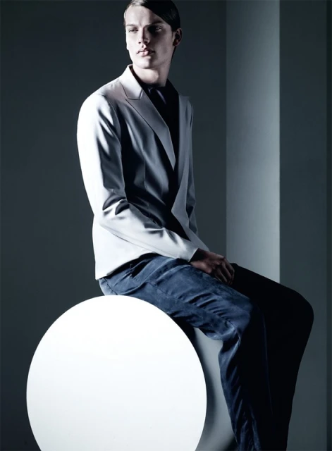 Fashion shot of a male model in a light grey suit sitting on a white sphere in a photo studio with directional lighting.