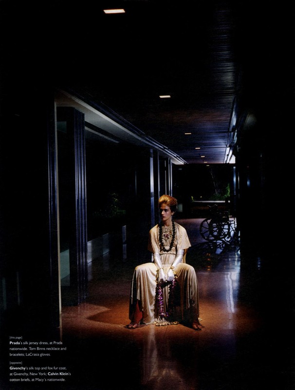 Indoor fashion shot features a lone model in a light dress and layered beaded necklaces, seated in a dimly lit hall with dark walls.