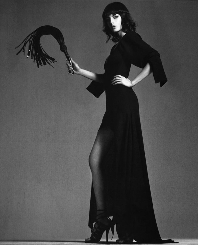 Monochrome fashion shot of a woman in a long black dress and cap, holding a whip, striking a pose against a grey backdrop.