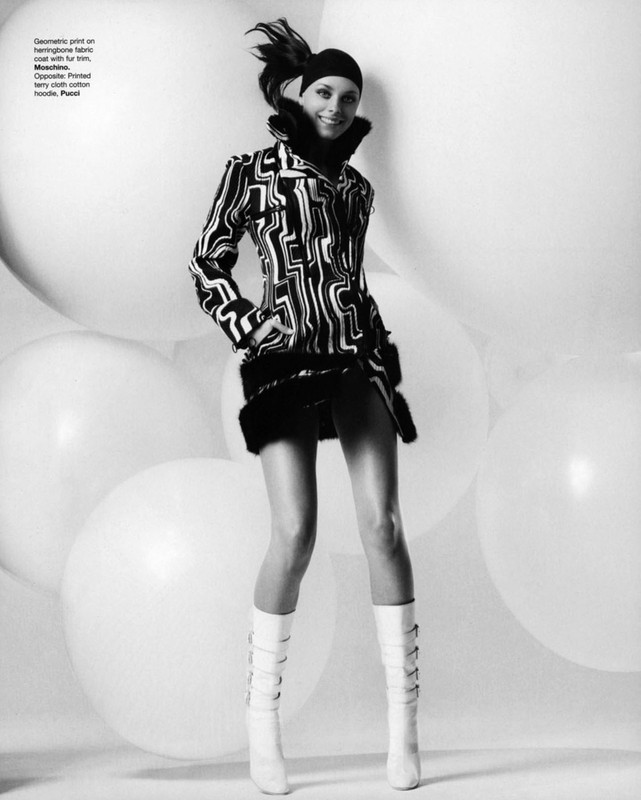 A stylish woman with a geometric coat poses in front of large white balloons, wearing white boots and a hat in a black and white fashion shot.