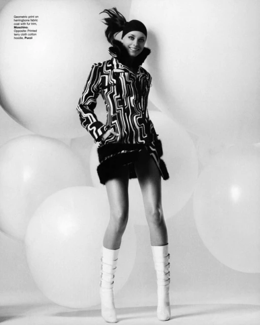 A stylish woman with a geometric coat poses in front of large white balloons, wearing white boots and a hat in a black and white fashion shot.
