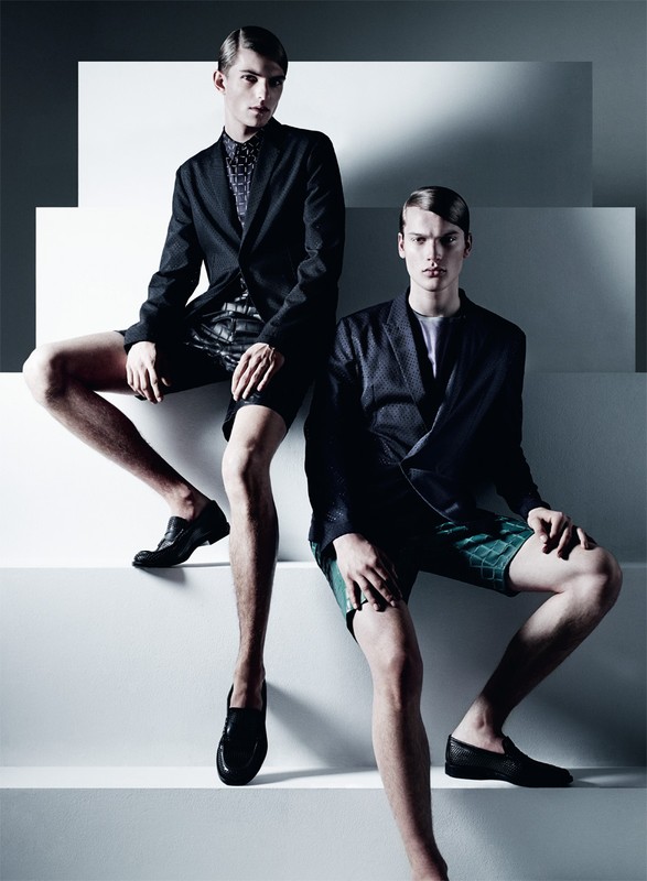 Two young male fashion models wearing blazers, shorts, and loafers pose on a white staircase, showcasing modern menswear against a light backdrop.