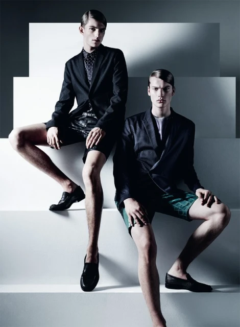 Two young male fashion models wearing blazers, shorts, and loafers pose on a white staircase, showcasing modern menswear against a light backdrop.