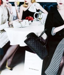 Three women seated around a white clothed restaurant table featuring luxury designer handbags and champagne glasses.