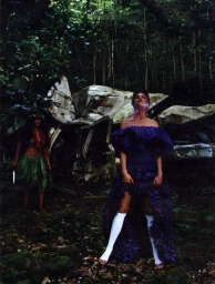 Woman in purple ruffled dress and white knee-high socks standing before a wrecked vehicle in a dense forest with another figure.