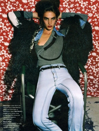 Model seated in a folding chair wearing light-wash jeans, a grey top, and a black feathered wrap against a red floral backdrop