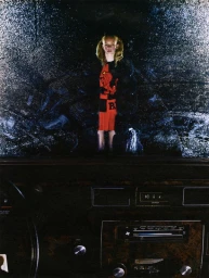 A young female model with blonde hair in pigtails wearing a red and black patterned dress reflected in a car windshield.
