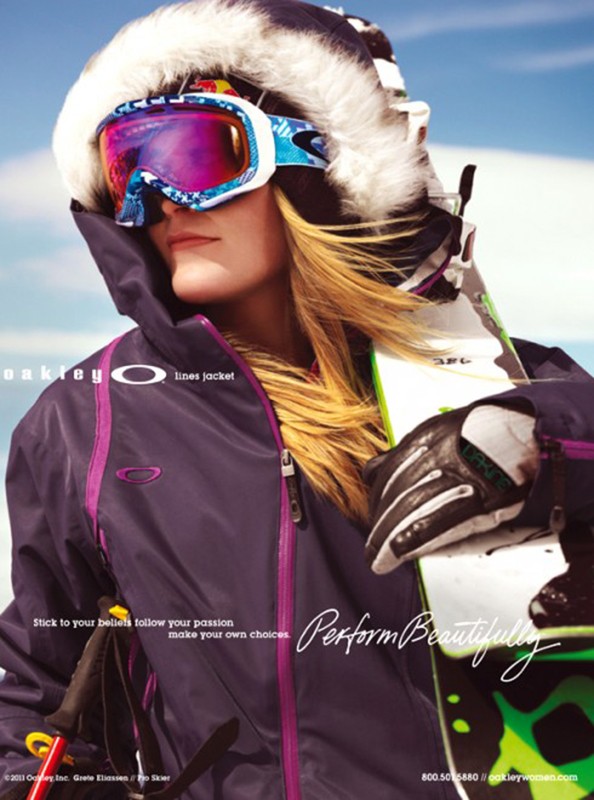 A woman models winter sportswear and ski goggles against a blue sky backdrop, showcasing outdoor fashion and athletic equipment.