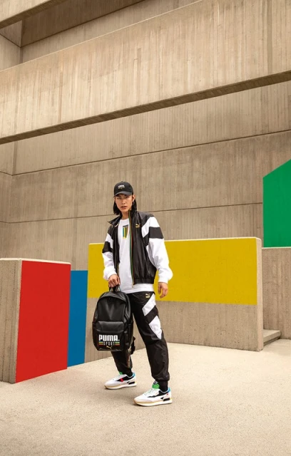 A person in Puma track suit and sneakers poses with a backpack against an abstract concrete and colourful wall background.
