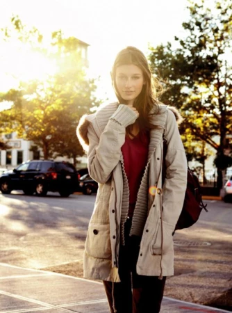 Young woman in a beige winter coat stands on a sunny city street, looking at the camera with trees and cars in the background.