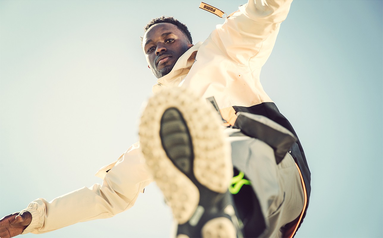 An African American man in a tan jacket with a Puma logo poses in a low-angle shot against a bright blue sky, showcasing fashion and athleticism.
