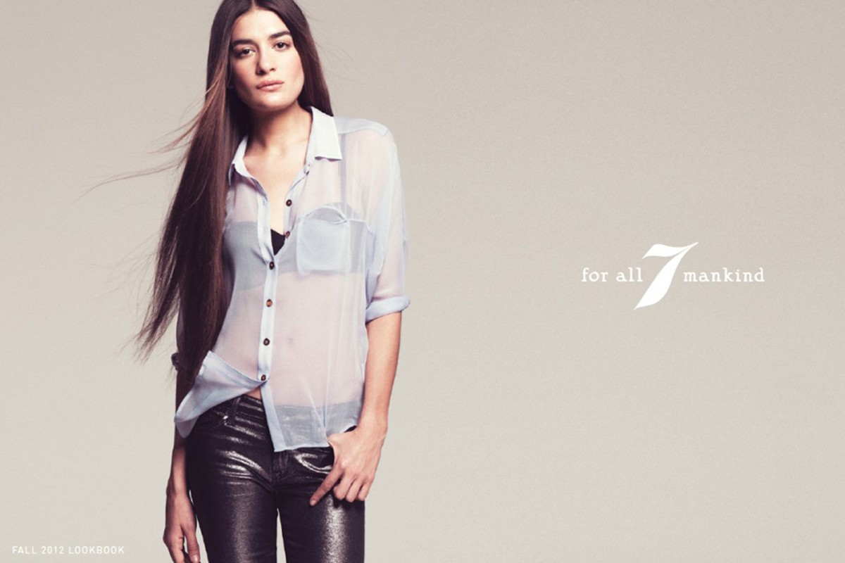 A woman with long brown hair poses in a sheer blue button-down shirt and shiny pants for a studio fashion portrait against a grey background.