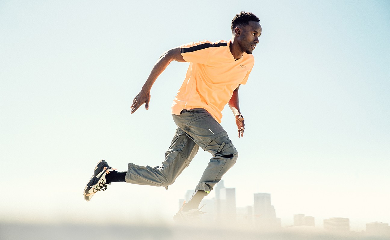 Athletic Black man in a peach t-shirt and grey cargo pants in motion, running with a city skyline in the background on a bright day.