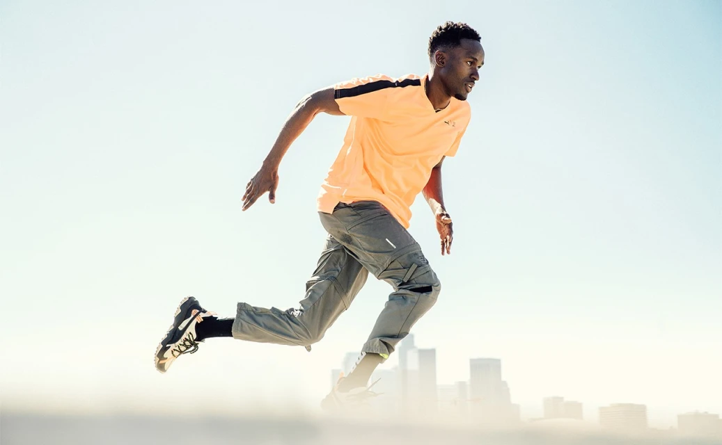 Athletic Black man in a peach t-shirt and grey cargo pants in motion, running with a city skyline in the background on a bright day.