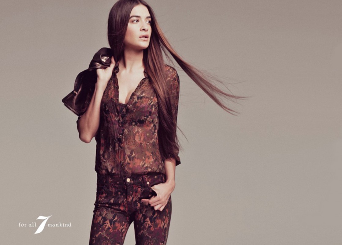 A brunette model in a patterned blouse and jeans holds a jacket in a fashion-style studio shot against a neutral gray backdrop.