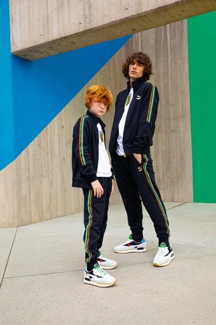 Two males, one adult and one child, wear matching black tracksuits with colorful stripes while posing outdoors against a concrete wall.