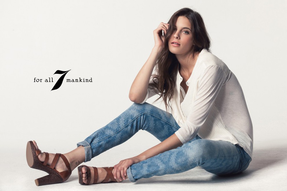 A woman with long brown hair sits on a white surface wearing a white shirt, blue jeans, and brown high-heeled sandals, with the logo visible.