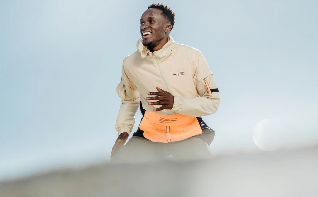 A smiling African-American man in a beige Puma jacket and bright orange belt, outdoor in daylight, with a clear sky backdrop.