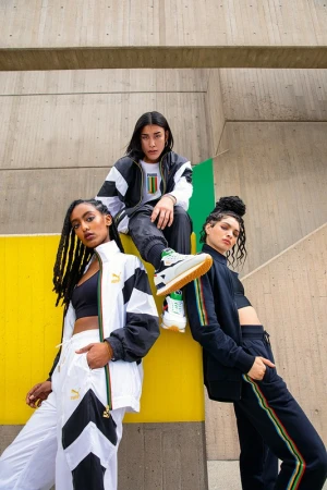 Three female models pose in Puma tracksuits in an urban setting. One woman sits above on a yellow and grey wall while the other two stand beside.