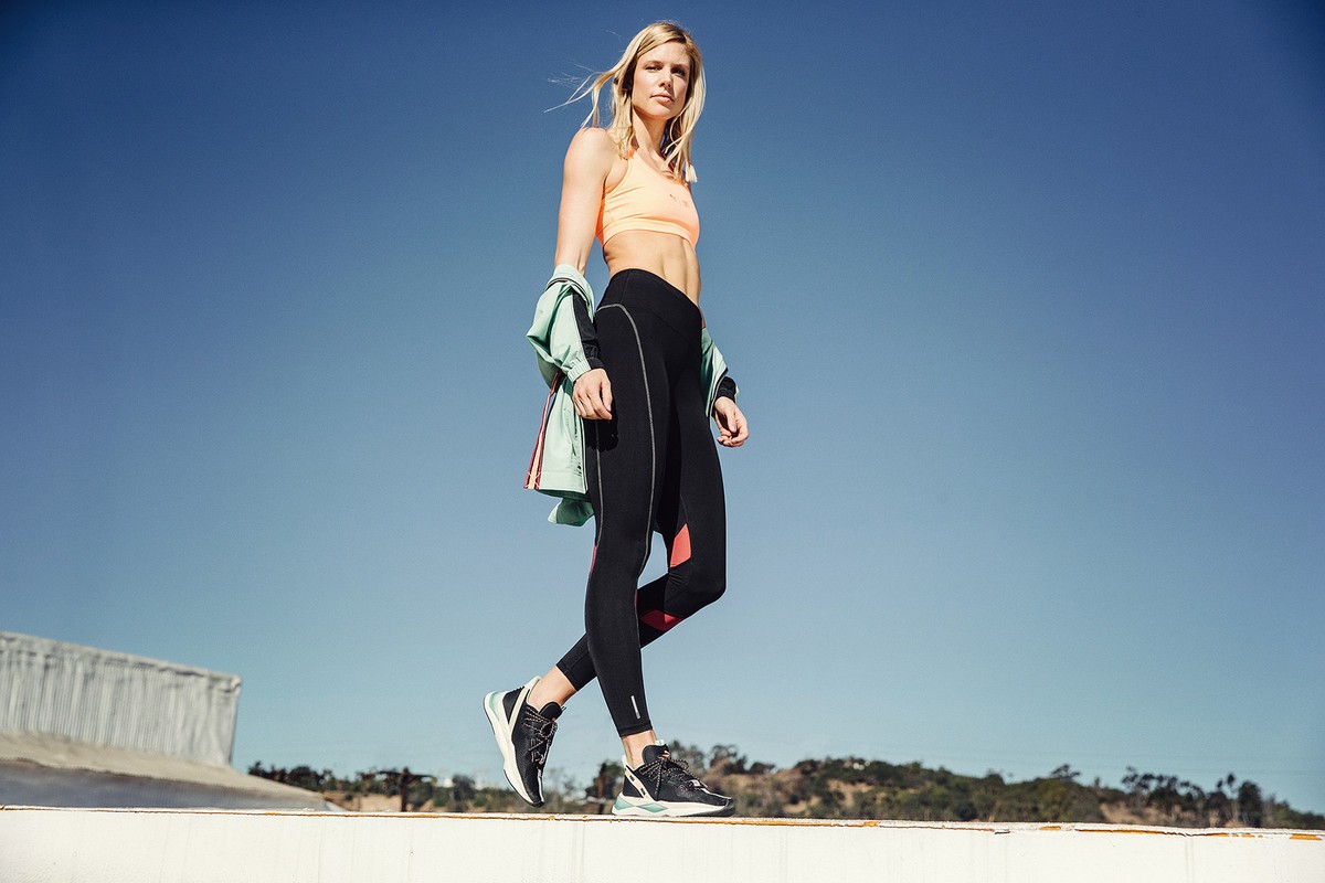 Athletic woman in workout clothes and black sneakers poses outdoors against a clear blue sky and a blurry landscape backdrop.