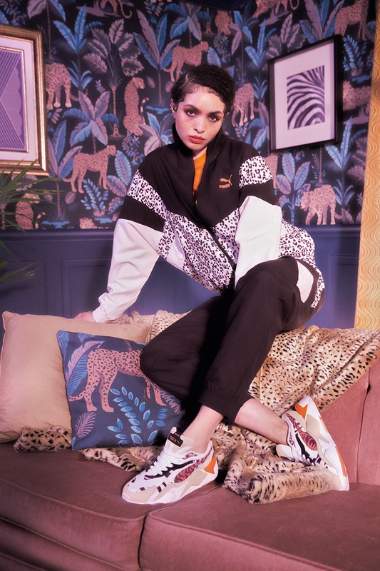 Woman in a Puma tracksuit sitting on a sofa with Puma sneakers, showcasing a blend of fashion and sportswear against a leafy backdrop.