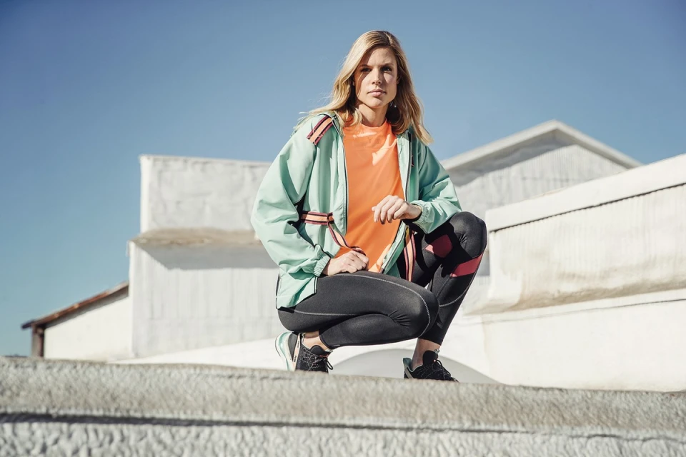 Female athlete in mint green jacket and black leggings squats on a concrete surface, set against a clear blue sky and minimalist white buildings.